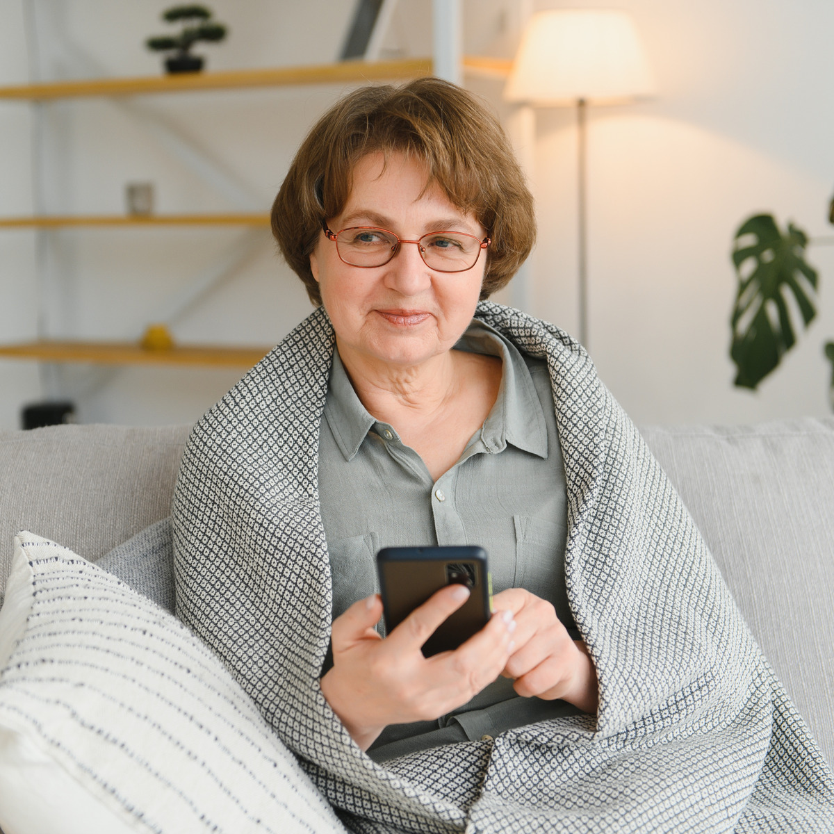 Ältere Frau sitzt lächelnd in eine Decke gehüllt auf dem Sofa und hält ein Smartphone in der Hand.
