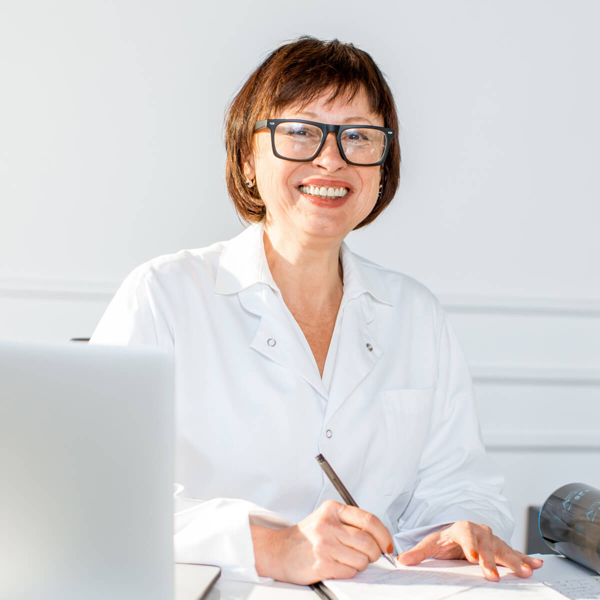 Frau sitzt an einem Schreibtisch vor einem Laptop, hält einen Stift und schaut lächelnd in die Kamera.